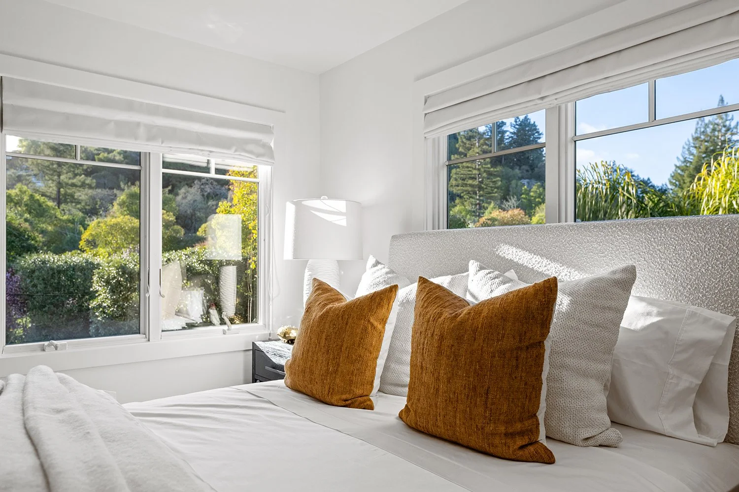 Bright bedroom with large windows showing greenery outside, white bed with white and orange pillows, and a white nightstand with a lamp.