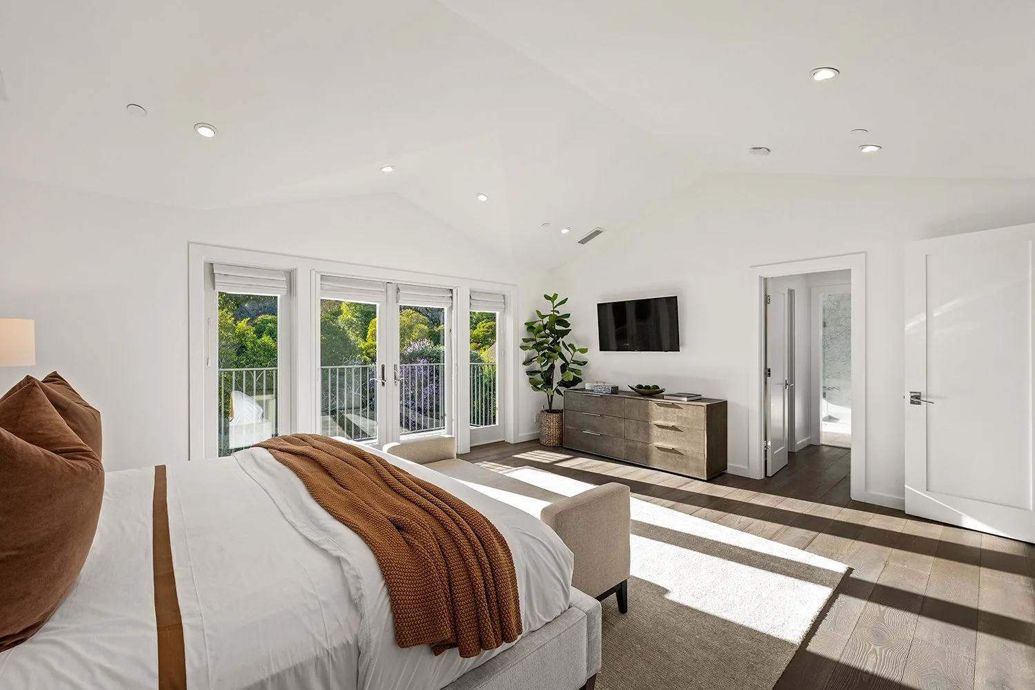 Bright bedroom with large windows, a bed with brown pillows and blanket, a beige armchair, a flat-screen TV on the wall, a gray dresser, a potted plant, and a balcony view of green trees.