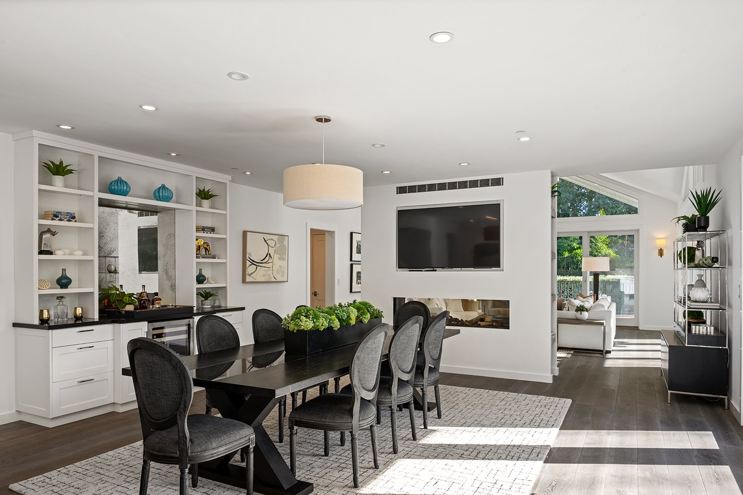 Modern open-concept living and dining room with white walls, dark wood floors, black dining table, gray upholstered chairs, built-in white shelves with decorative items, large TV mounted on wall, fireplace, and large windows with greenery outside.