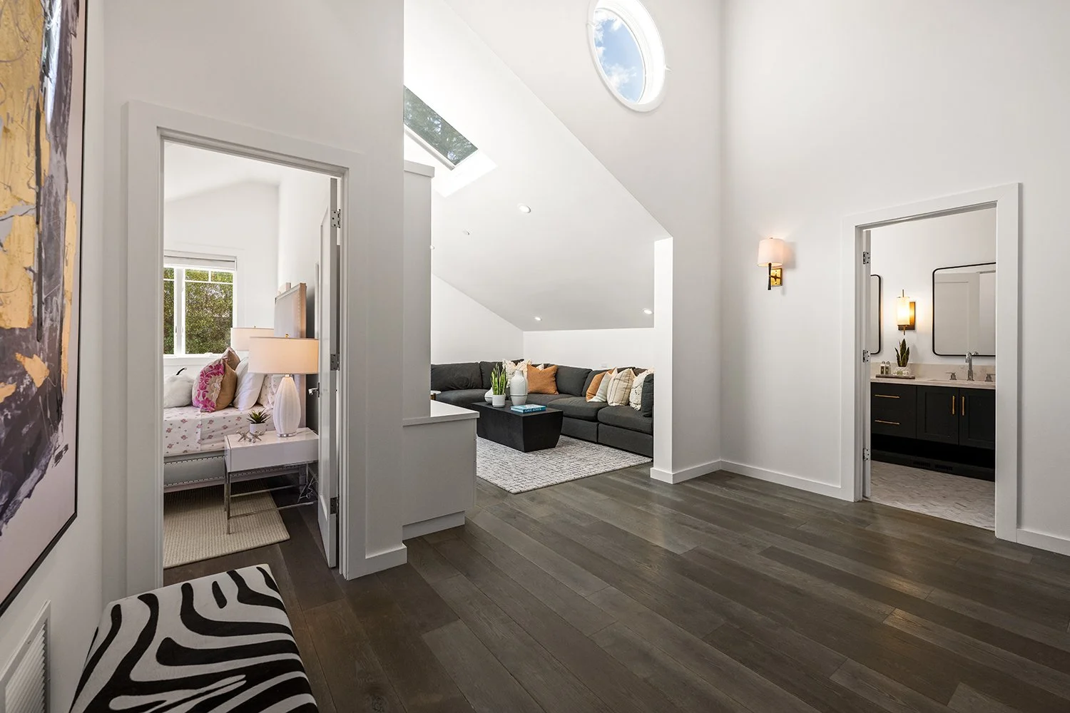 Modern living space with white walls, dark wood flooring, a black sectional sofa with throw pillows, a coffee table, and a small bedroom with a bed, lamp, and window visible through open doors. A bathroom with black vanity and mirrors is also visible