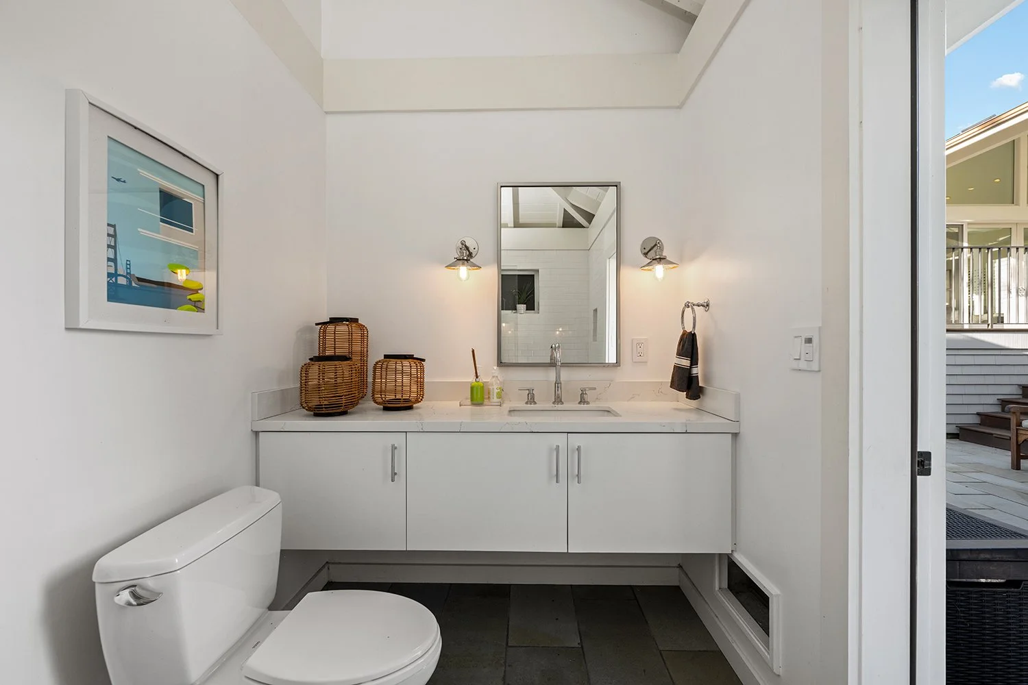 A modern bathroom with white walls, a white vanity with a marble countertop, a rectangular mirror, two wall-mounted lights, a wicker basket, a green soap dispenser, and a towel on a ring. A white toilet is visible on the left, and an open door reveal