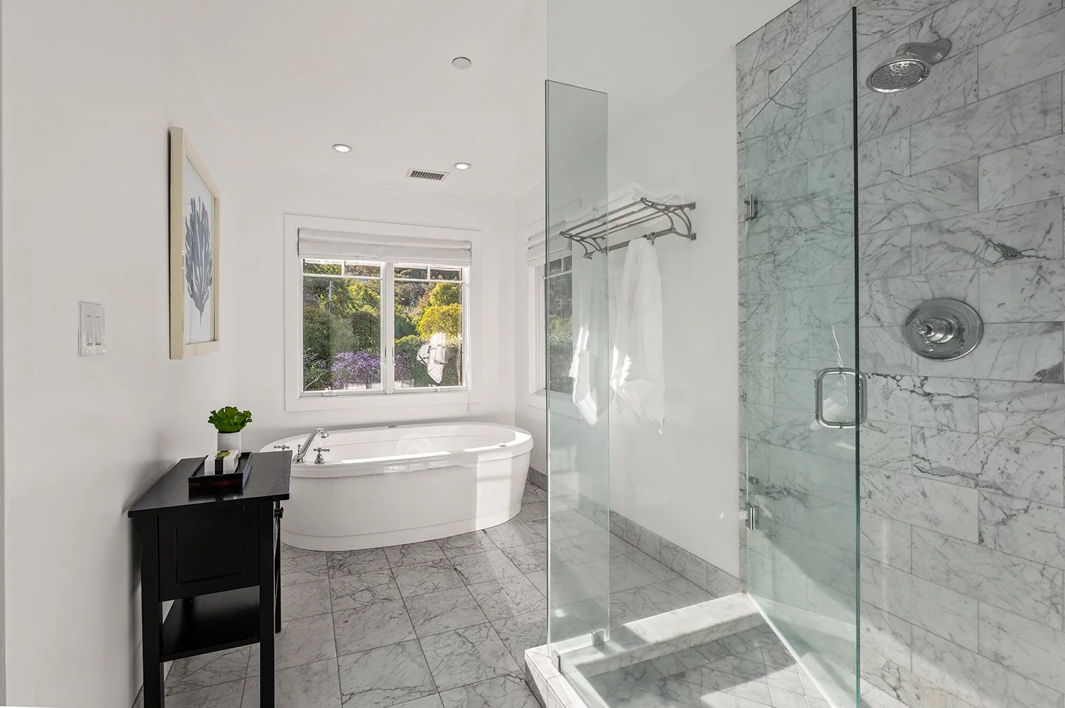 Modern bathroom with marble tiles, a glass shower, a white bathtub, and a window showing greenery outside.
