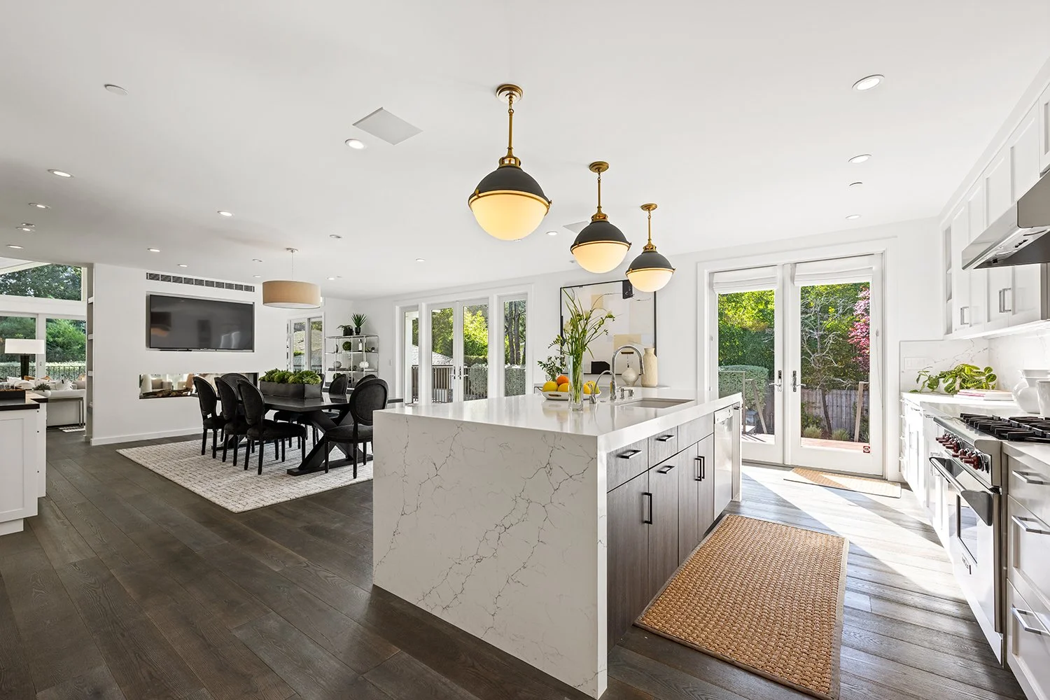 Bright, modern kitchen with white cabinetry, a large island with marble countertop, pendant lighting, and a view of a dining area with black chairs and a wall-mounted TV.