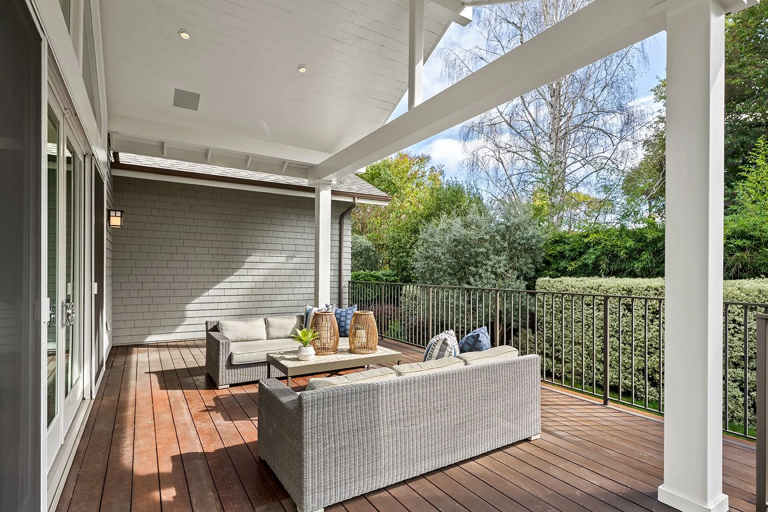 An outdoor deck with wicker sofas, a white brick wall, potted plants, lanterns, and surrounded by trees and greenery under a partly cloudy sky.