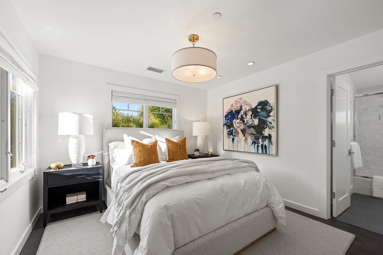 Bright bedroom with white walls, a large bed with white bedding and two brown pillows, black nightstands with lamps, and an abstract painting on the wall. A window shows greenery outside, and an open door reveals a bathroom.