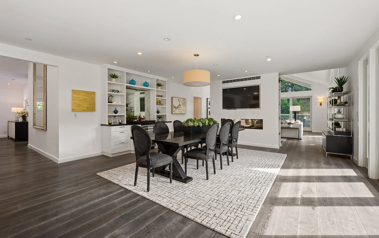 Modern dining room with a black table and upholstered chairs, a large wall-mounted TV, built-in shelving with decor, and a living area with glass doors leading outside.