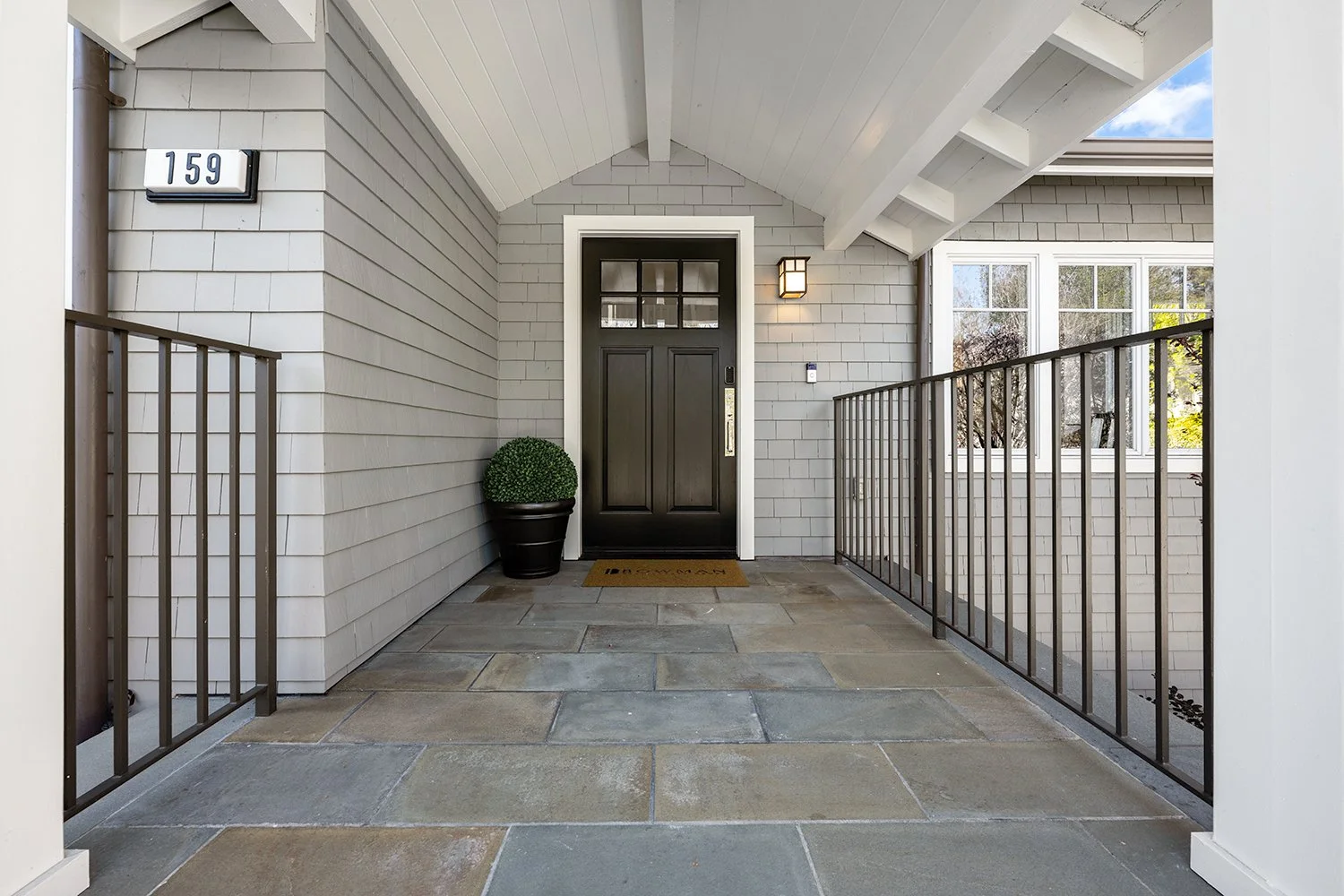 Front porch of a house with a black front door, potted plant, and a black railing, with house number 159 on the wall.