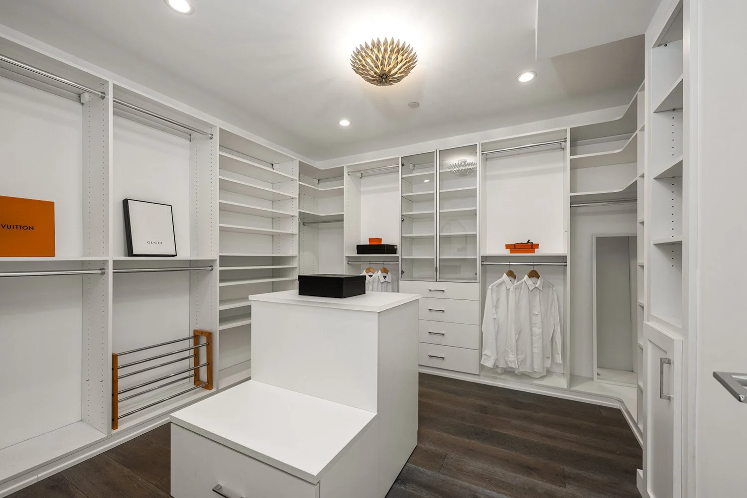 Empty walk-in closet with white shelving units, hanging rods, a central island, some boxes, and a few hanging shirts, with a decorative ceiling light.
