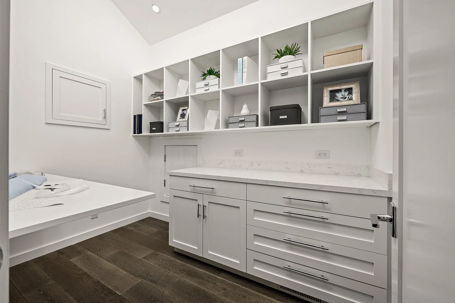 A white bedroom with a built-in shelf filled with decorative items, plants, photo frames, and storage boxes. There is a white cabinet with drawers and a marble countertop below the shelf, and a bed with white bedding and light blue pillows to the sid