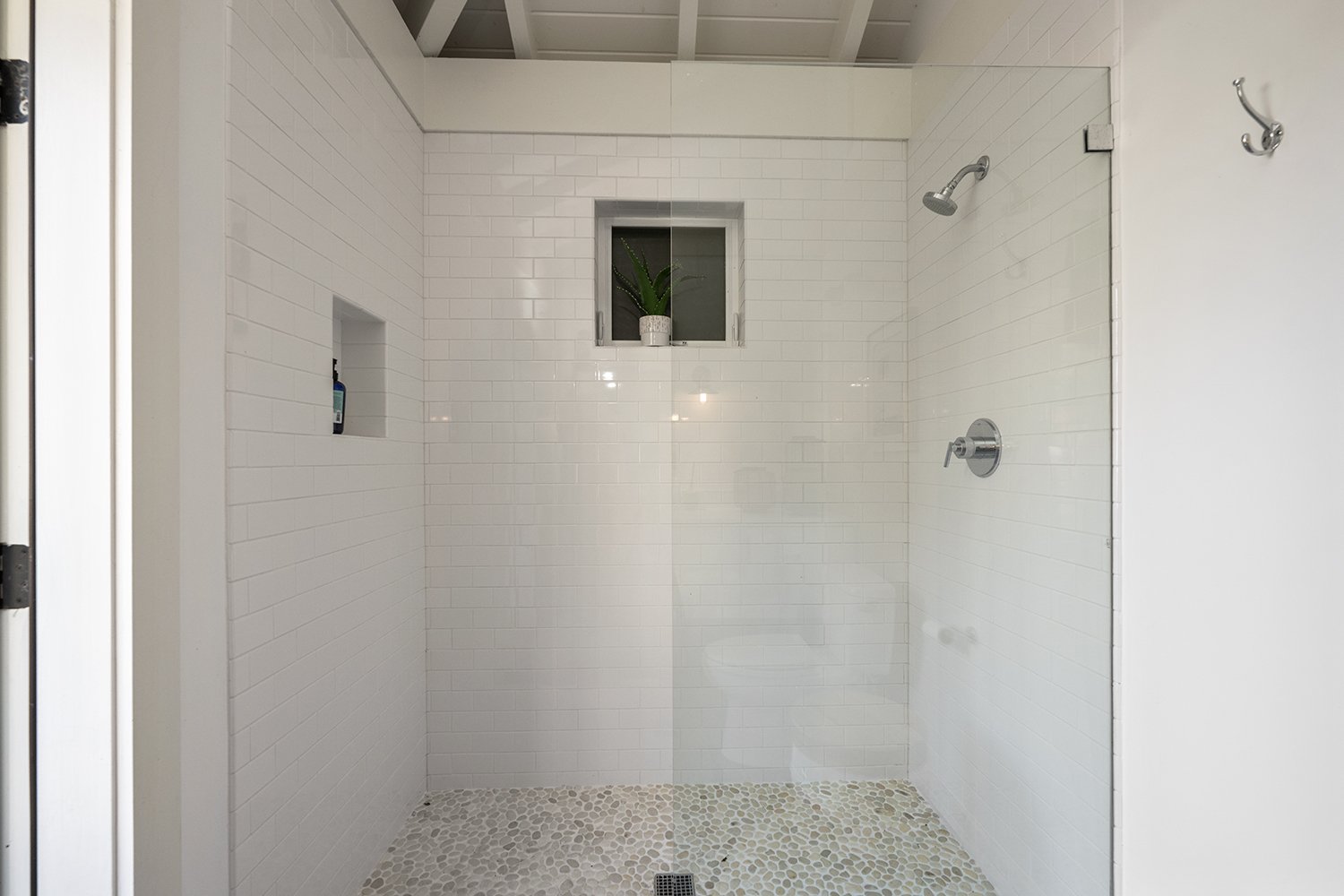 Empty walk-in shower with white subway tile walls, a small window with a potted plant, and pebble flooring.