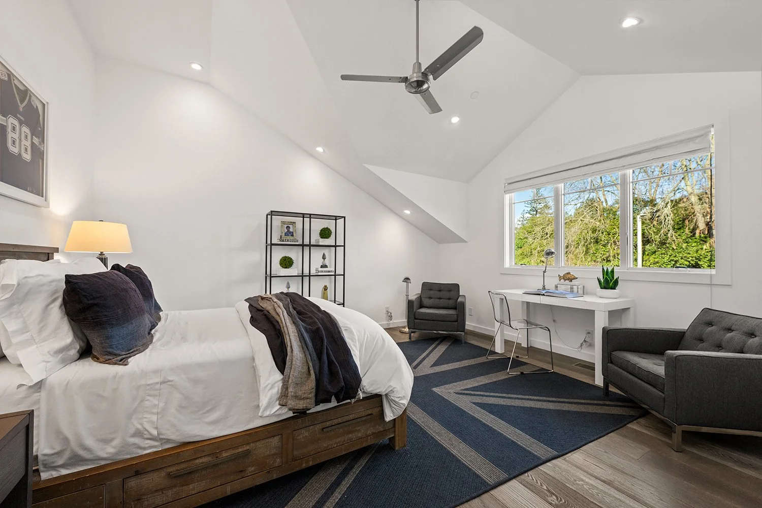 Spacious bedroom with a large bed, dark pillows, and a throw blanket, two armchairs, a black metal shelf with decorative items, a white desk with a chair and a plant, large window with a view of trees, ceiling fan, and ceiling lights.