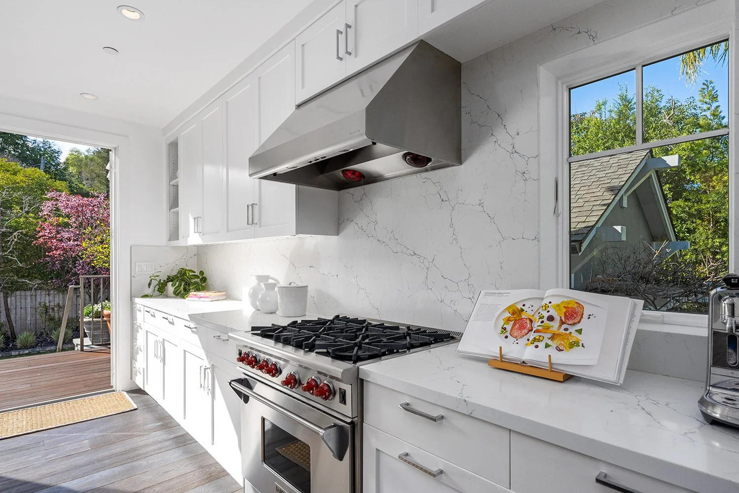 Modern white kitchen with marble countertops, stainless steel stove, and a cookbook open on a stand. Large windows reveal a backyard with trees and a pink flowering tree.