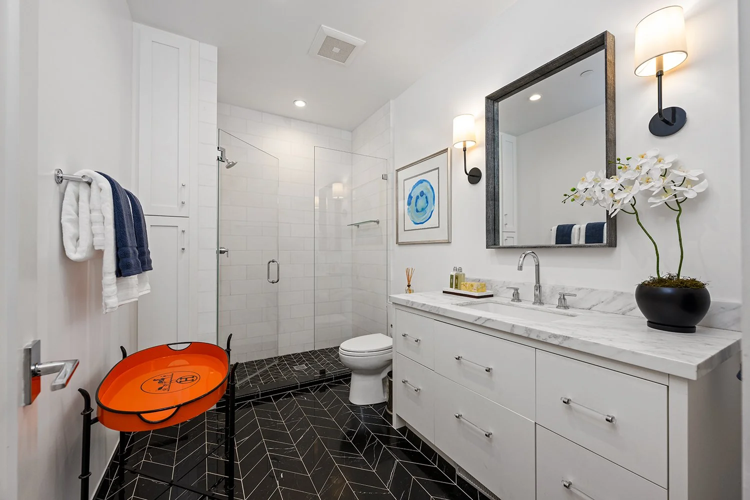 Modern white bathroom with black geometric floor tiles, a shower area with white subway tiles and glass door, a white vanity with marble countertop, a large mirror, and decorative items including a potted orchid and framed art.