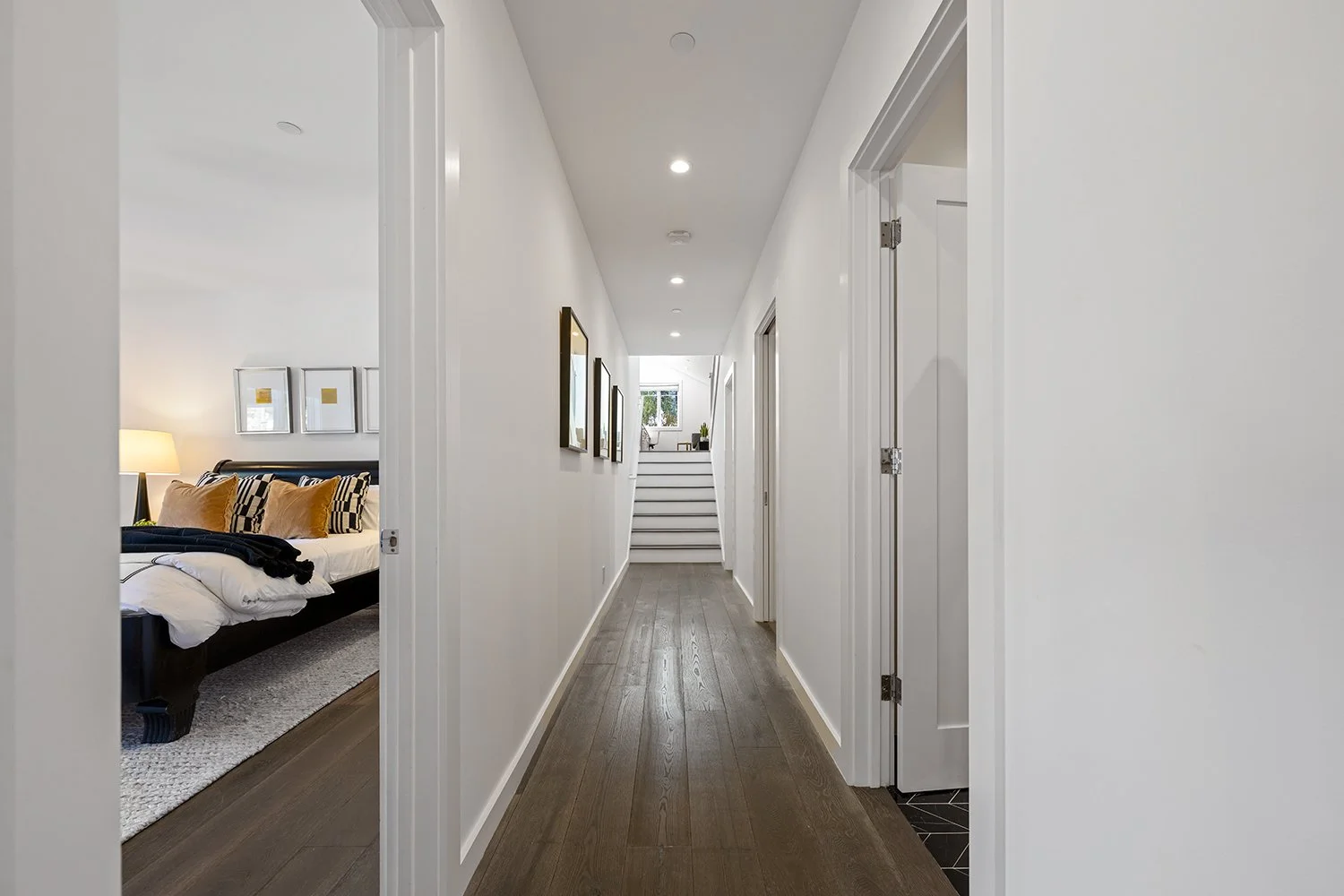 A bright hallway with wood flooring, white walls, and ceiling-mounted recessed lighting. Artworks line the left wall, leading to a staircase at the end, which is visible through a window at the top with some greenery outside.