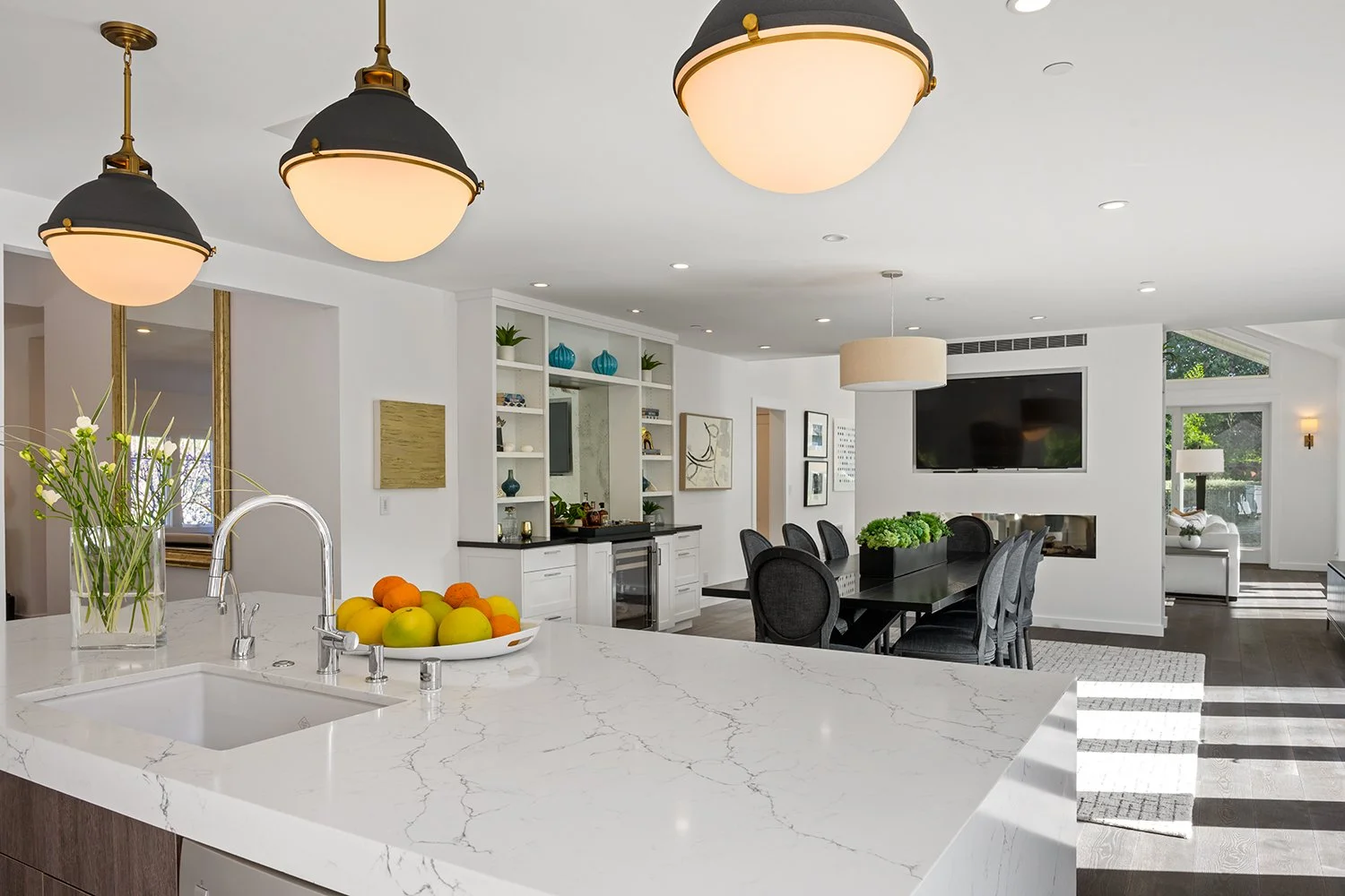 Modern open-concept kitchen and dining area with white marble island, black dining table with gray chairs, and built-in shelving with decorative items, a large TV, and a cozy living space in the background.