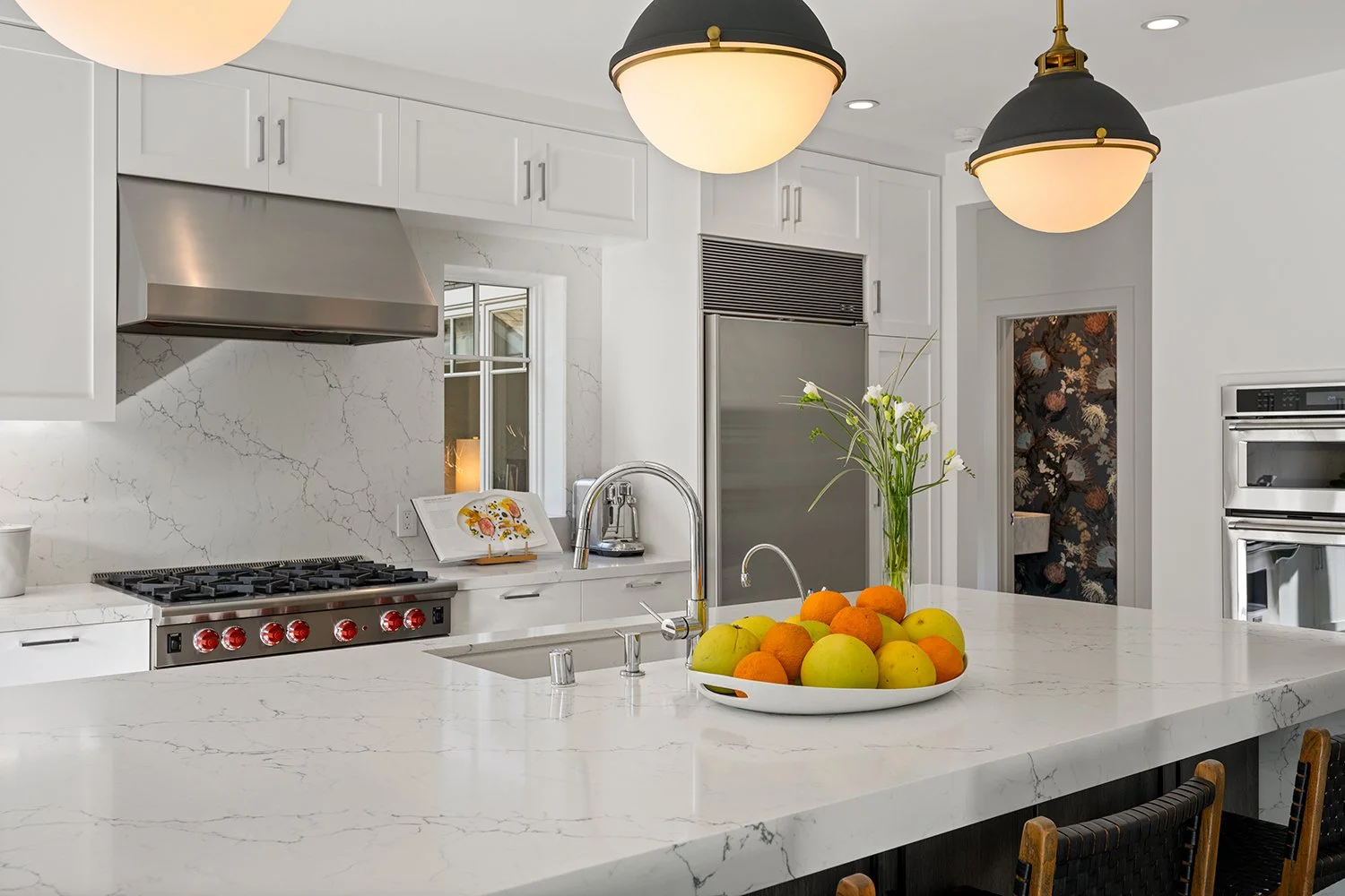 Modern white kitchen with marble countertops, stainless steel appliances, pendant lights, and a large bowl of apples and oranges on the island.