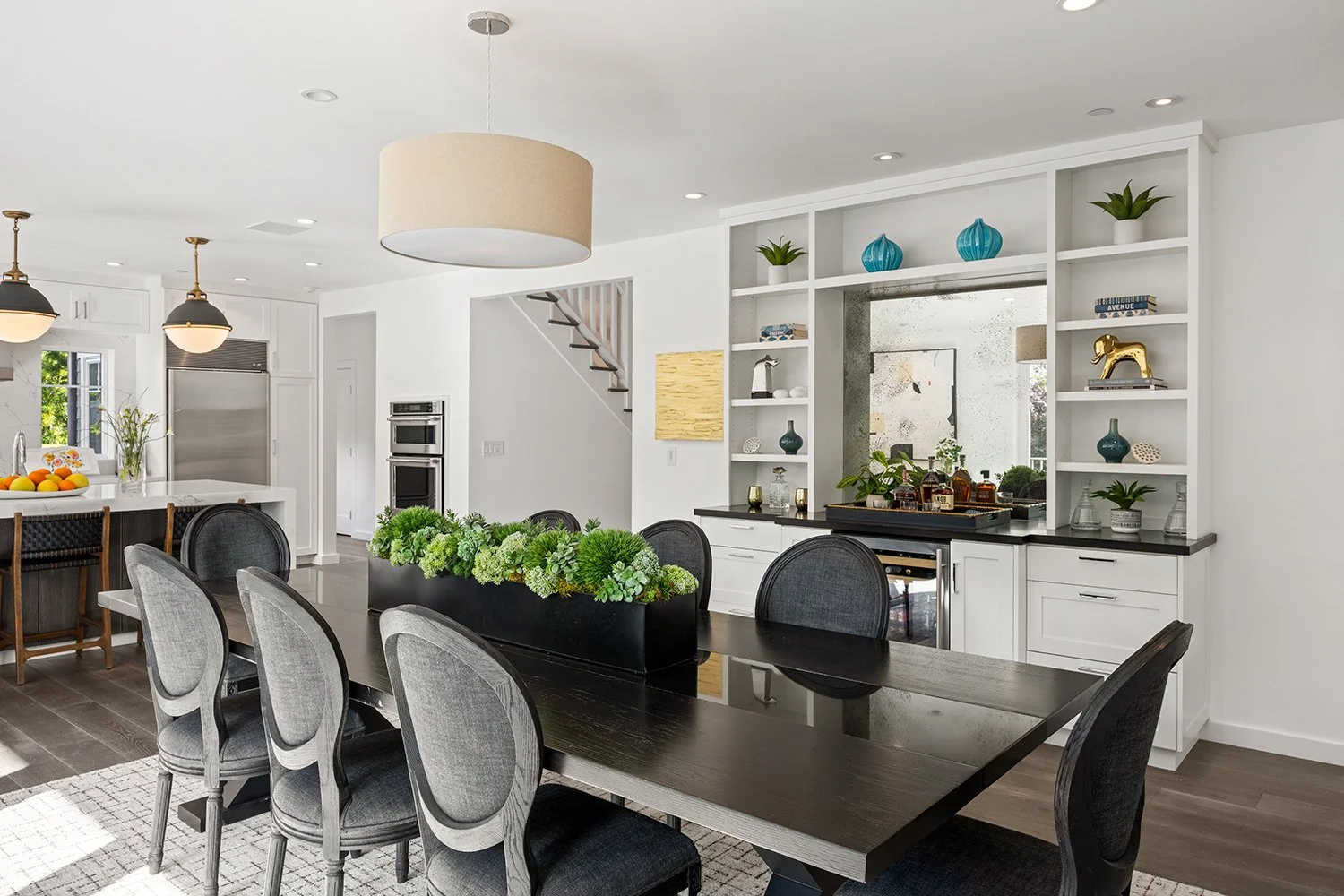 Modern dining room with dark wood table, gray chairs, white built-in shelving, and decorative items including plants and vases, adjacent to a kitchen with white cabinets and island.