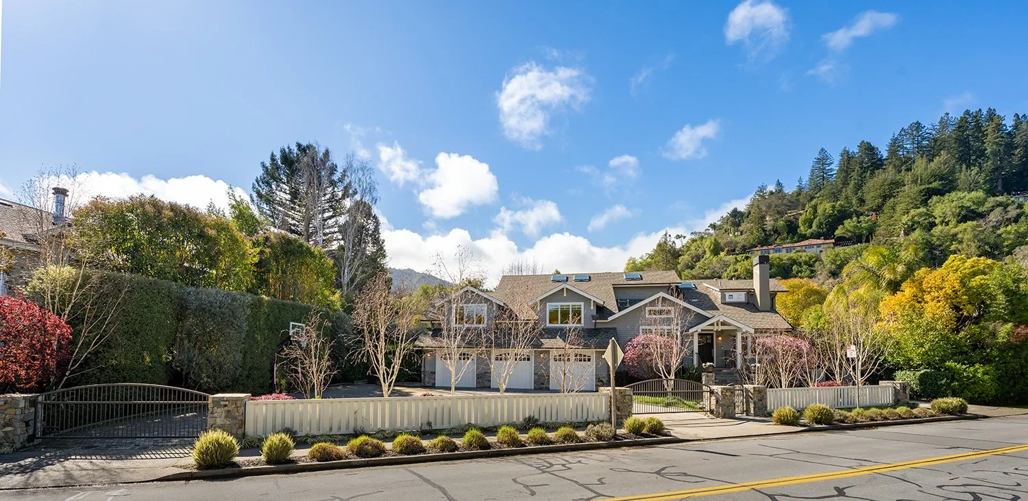 Large house with a front yard, trees, and a driveway, set against a hillside with lush greenery and a partly cloudy sky.