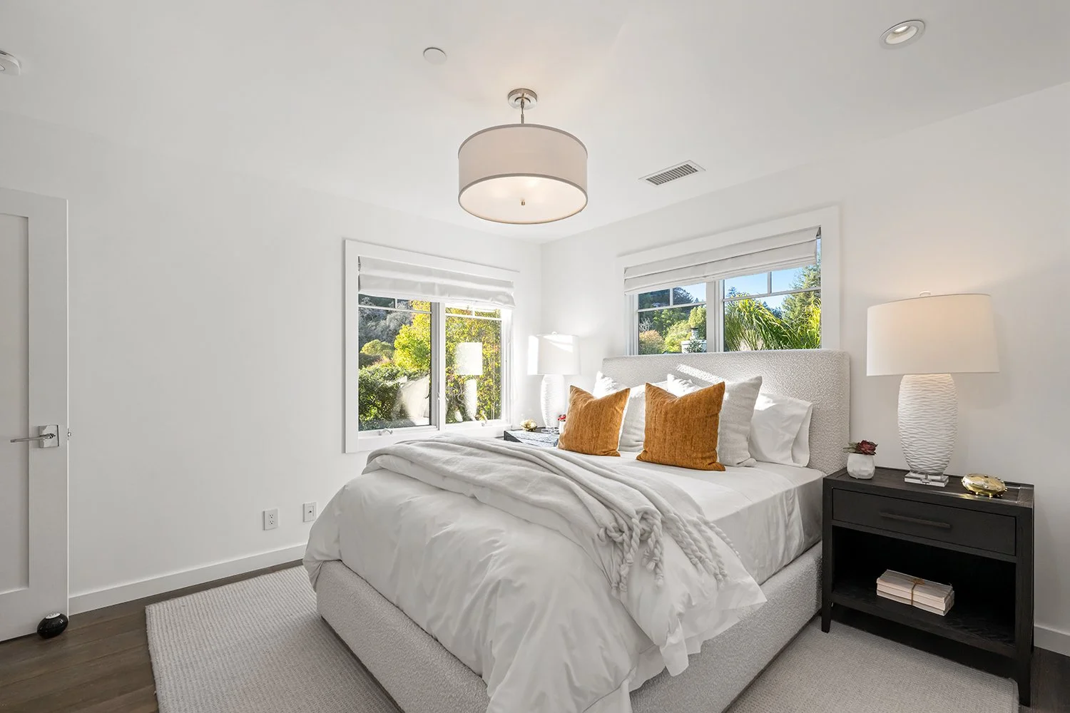 Bright bedroom with white walls, a bed with white linens and mustard-colored pillows, a black nightstand with a white lamp, and large windows showing greenery outside.