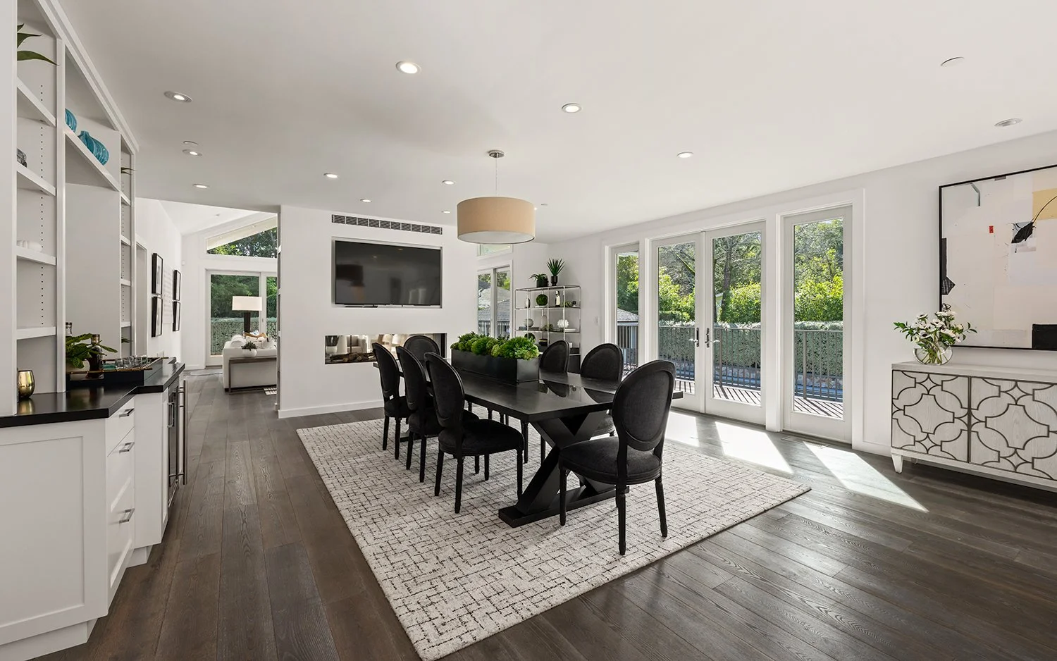 Modern dining room with black table and eight black chairs, large window doors, white walls, hardwood floor, light fixture, artwork, and a view of green trees outside.