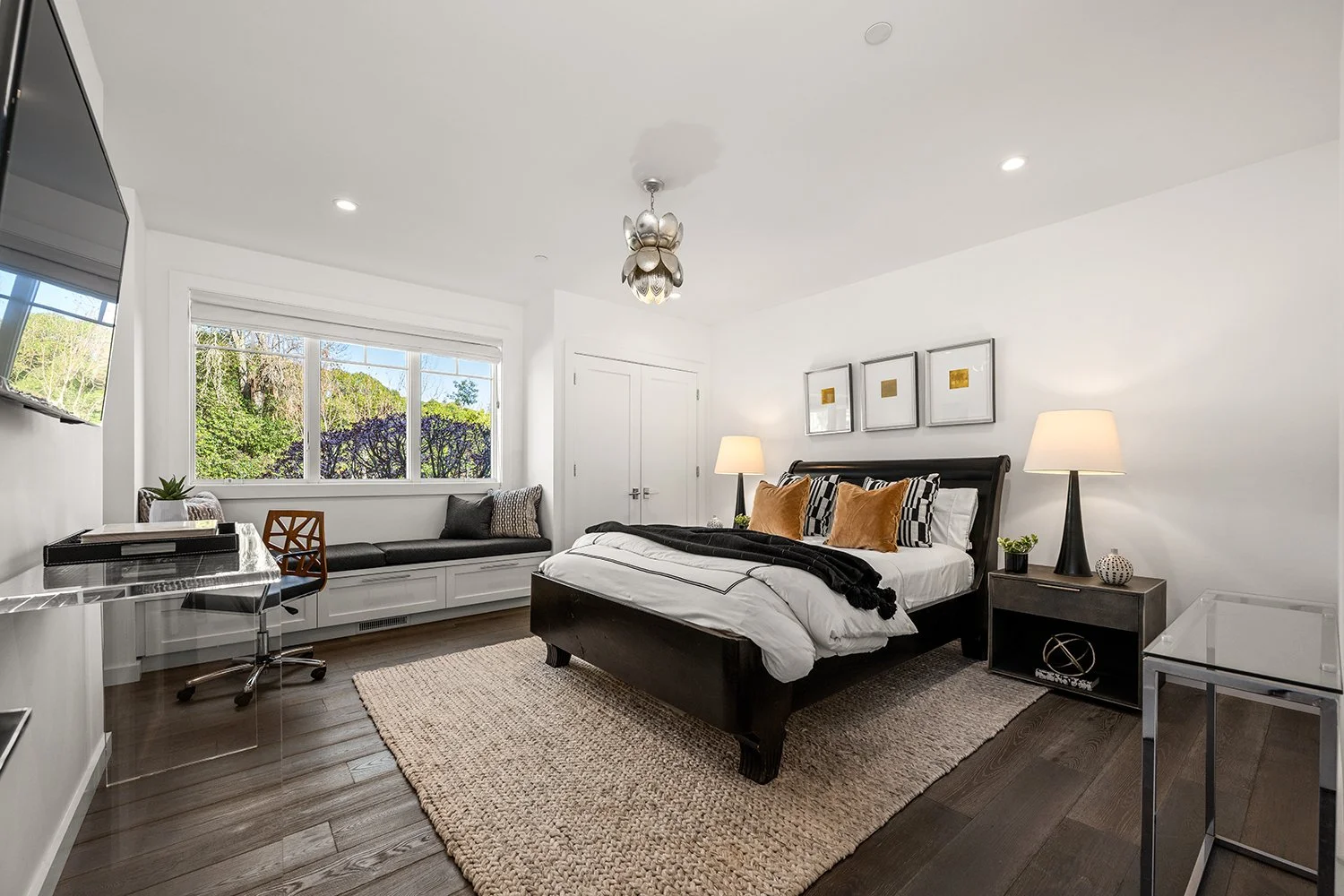 Modern bedroom with black bed frame, white bedding, and decorative pillows, two bedside lamps, a window seat with pillows, a window with greenery outside, and a wall-mounted TV.