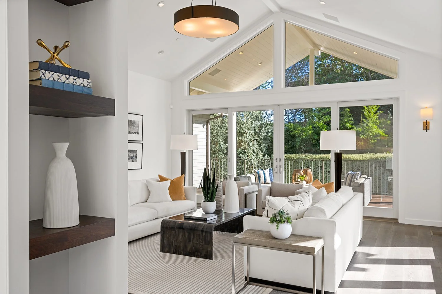 Bright living room with large windows, white sofas, wooden and black coffee tables, potted plants, and a view of a backyard with trees and a deck.