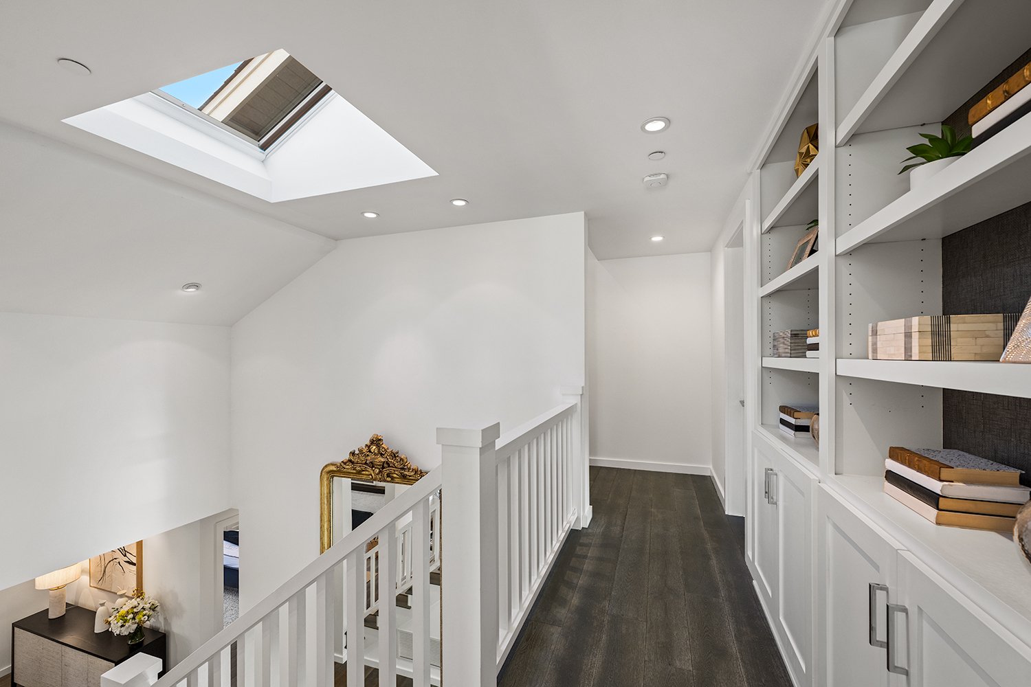 Indoor hallway with white walls, dark wooden flooring, a built-in white bookshelf on the right, a skylight window on the ceiling, and a decorative mirror with a gold ornate frame on the staircase wall.