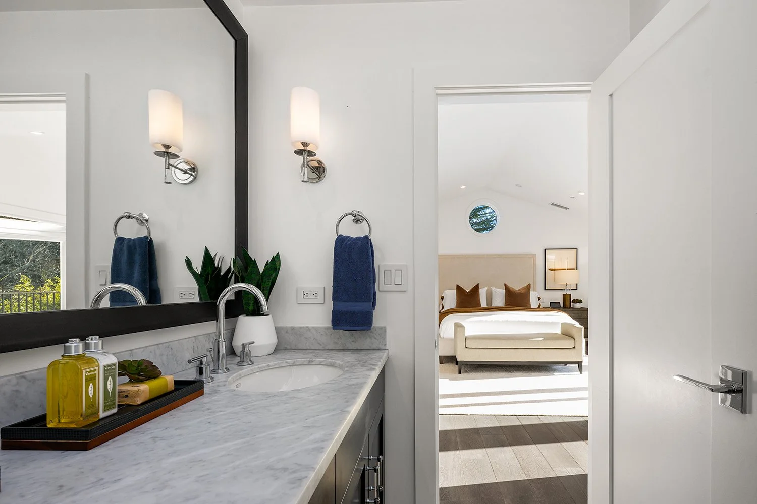 Bathroom with marble countertop, plant, yellow and green soap bottles, large mirror, and wall-mounted lights, leading to a bedroom with a beige bed, pillows, and a round window.
