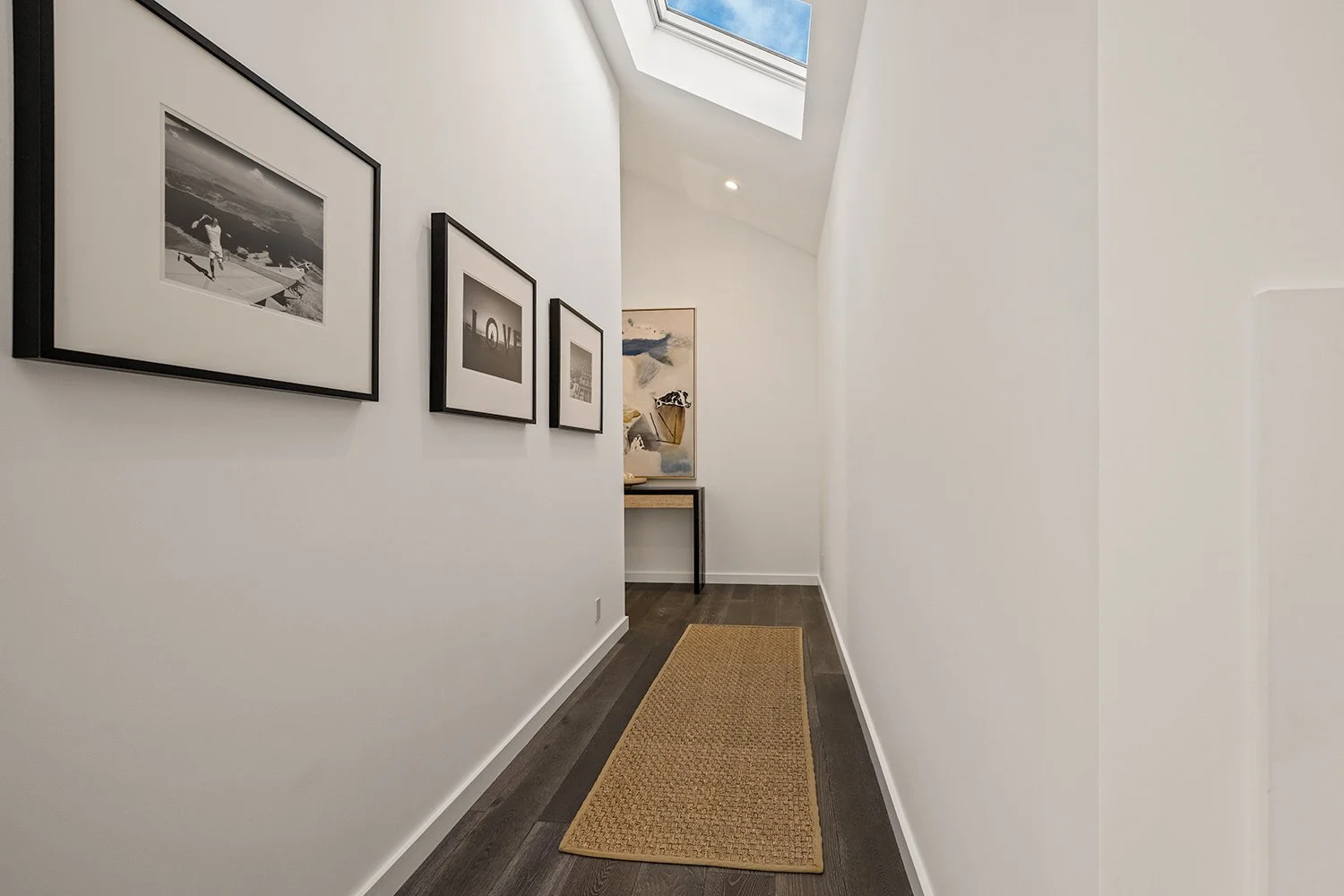 A narrow hallway with white walls, dark wood flooring, a beige rug, and framed black-and-white photographs. A skylight overhead allows natural light to enter.