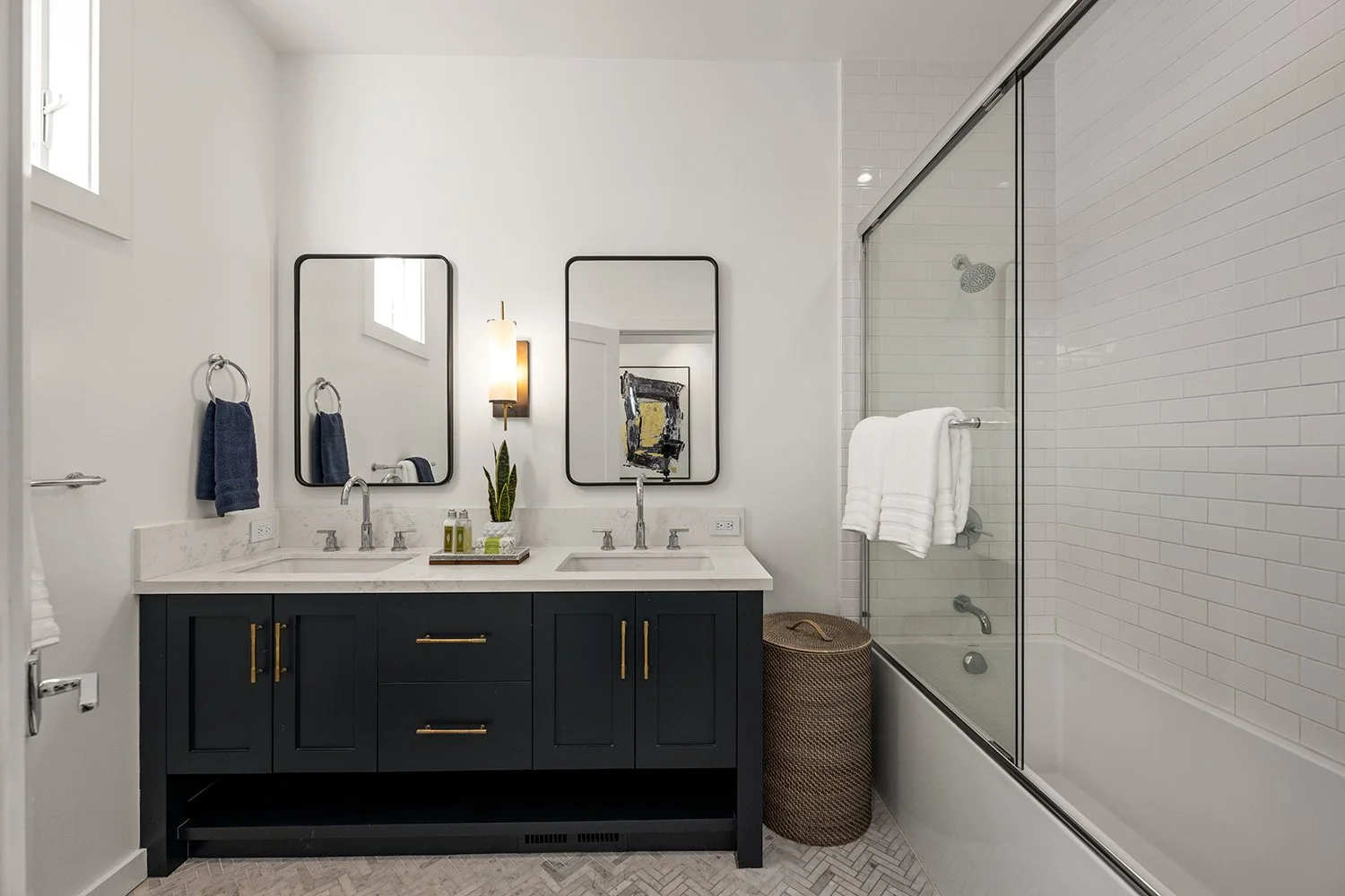 Modern bathroom with black vanity, double sinks, two mirrors, wall-mounted light, wicker laundry basket, towel rack with white towels, shower with glass door, white subway tile, and blue towels.