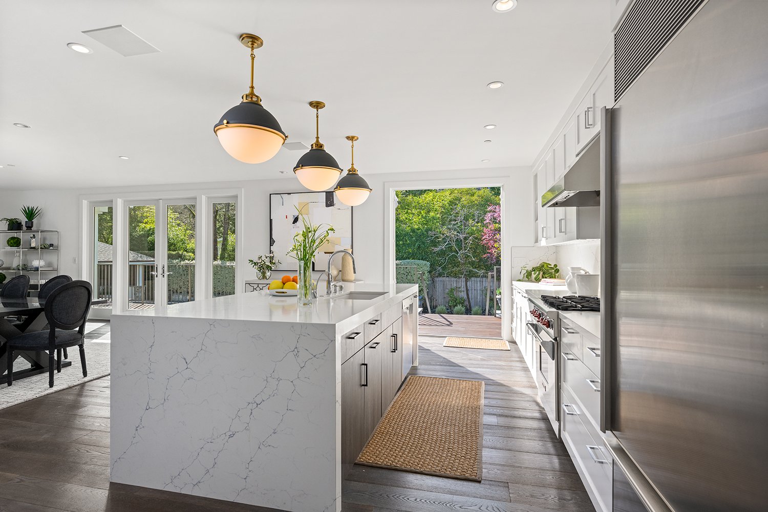 Modern kitchen with white marble island, stainless steel refrigerator, white cabinetry, black pendant lights, and a view of a backyard with trees and a deck.