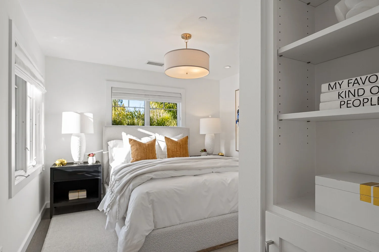 Bright bedroom with a white bed, two orange pillows, two white lamps, and a window showing trees outside.