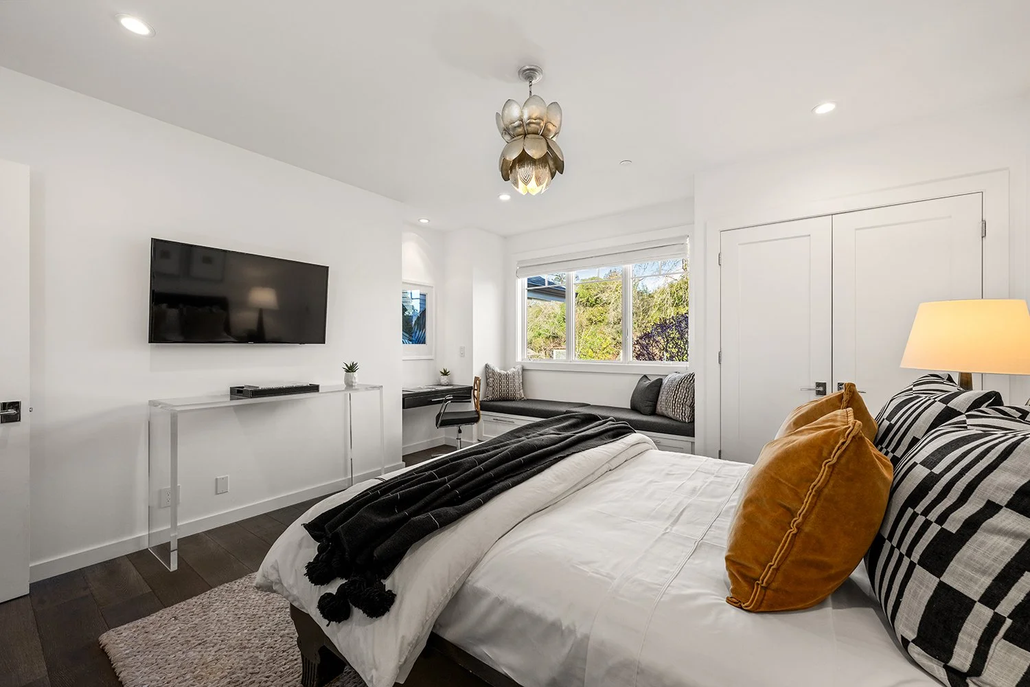 A modern bedroom with a large bed featuring plaid and velvet pillows, a white duvet, and a black throw. There's a wall-mounted flat-screen TV on the white wall and a window seat with cushions by the window. The room has a small desk with a chair unde