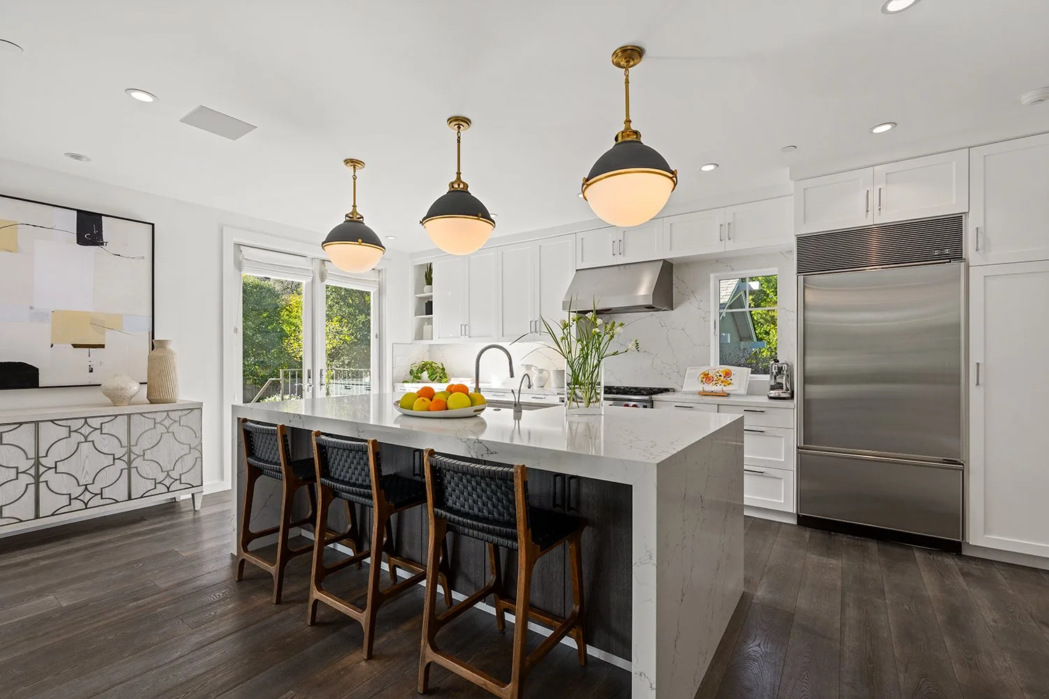 Modern kitchen with white cabinets, a large island with a marble countertop, and pendant lighting. There are three black bar stools at the island, stainless steel appliances, and windows with a view of greenery outside.