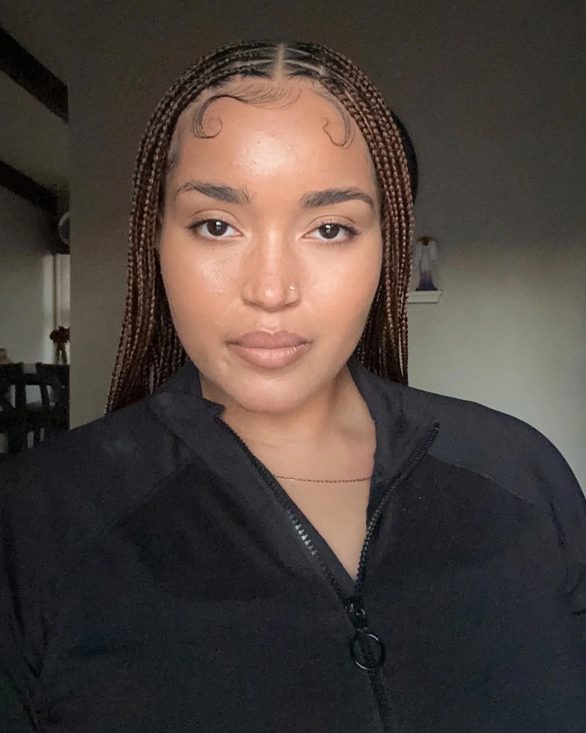 A young woman with braided hair styled with curls at the front, wearing a black zip-up jacket and a thin necklace, taking a selfie indoors with a neutral background.