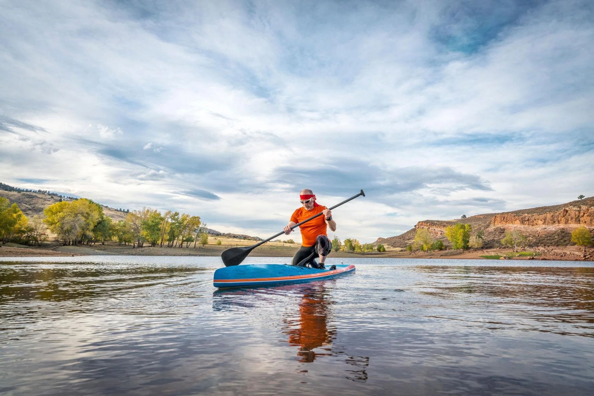 Person paddleboarding on a calm lake with trees and hills in the background under a partly cloudy sky.