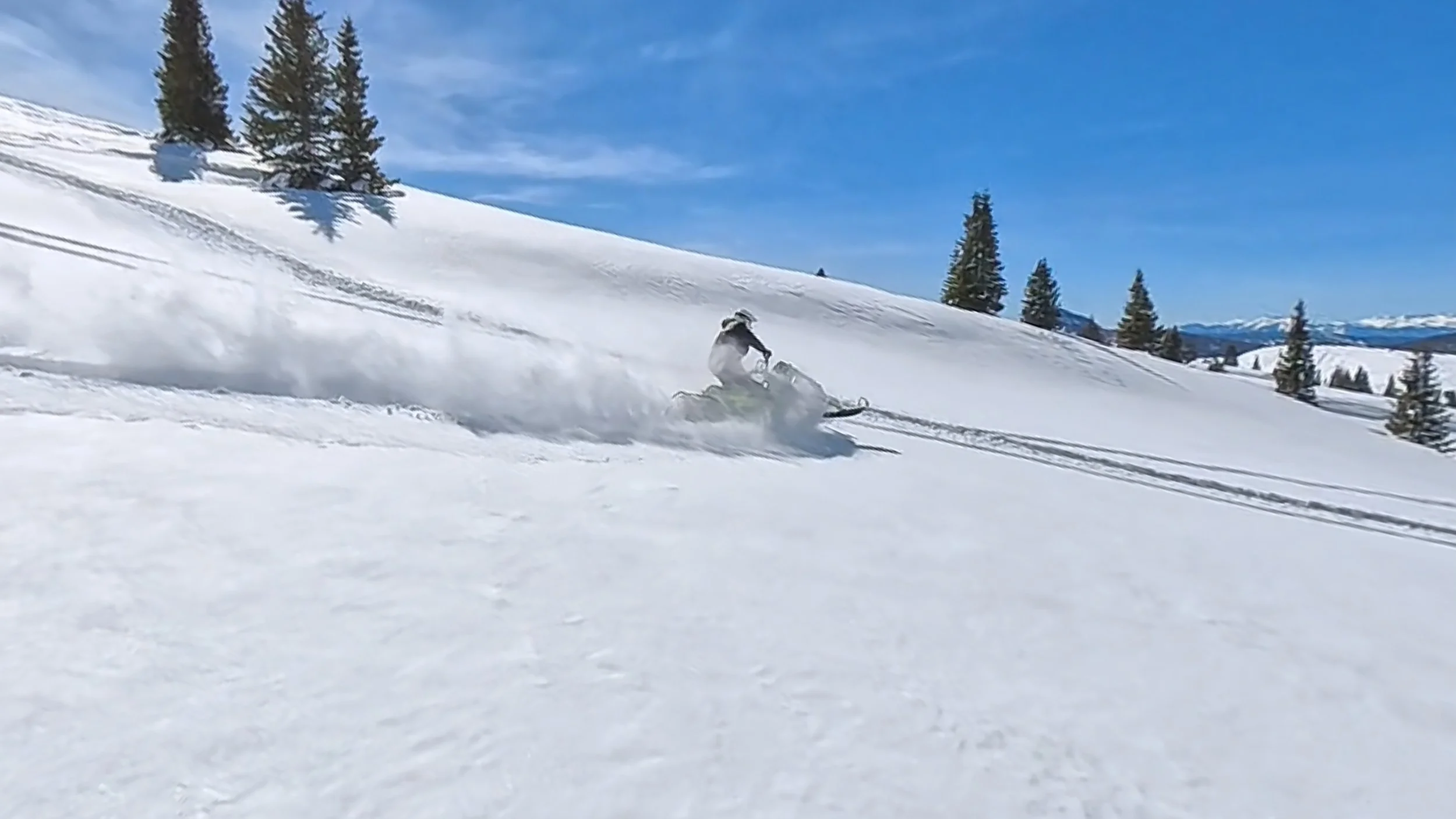 Person snowmobiling in a snowy landscape with pine trees and mountains in the background under a blue sky.