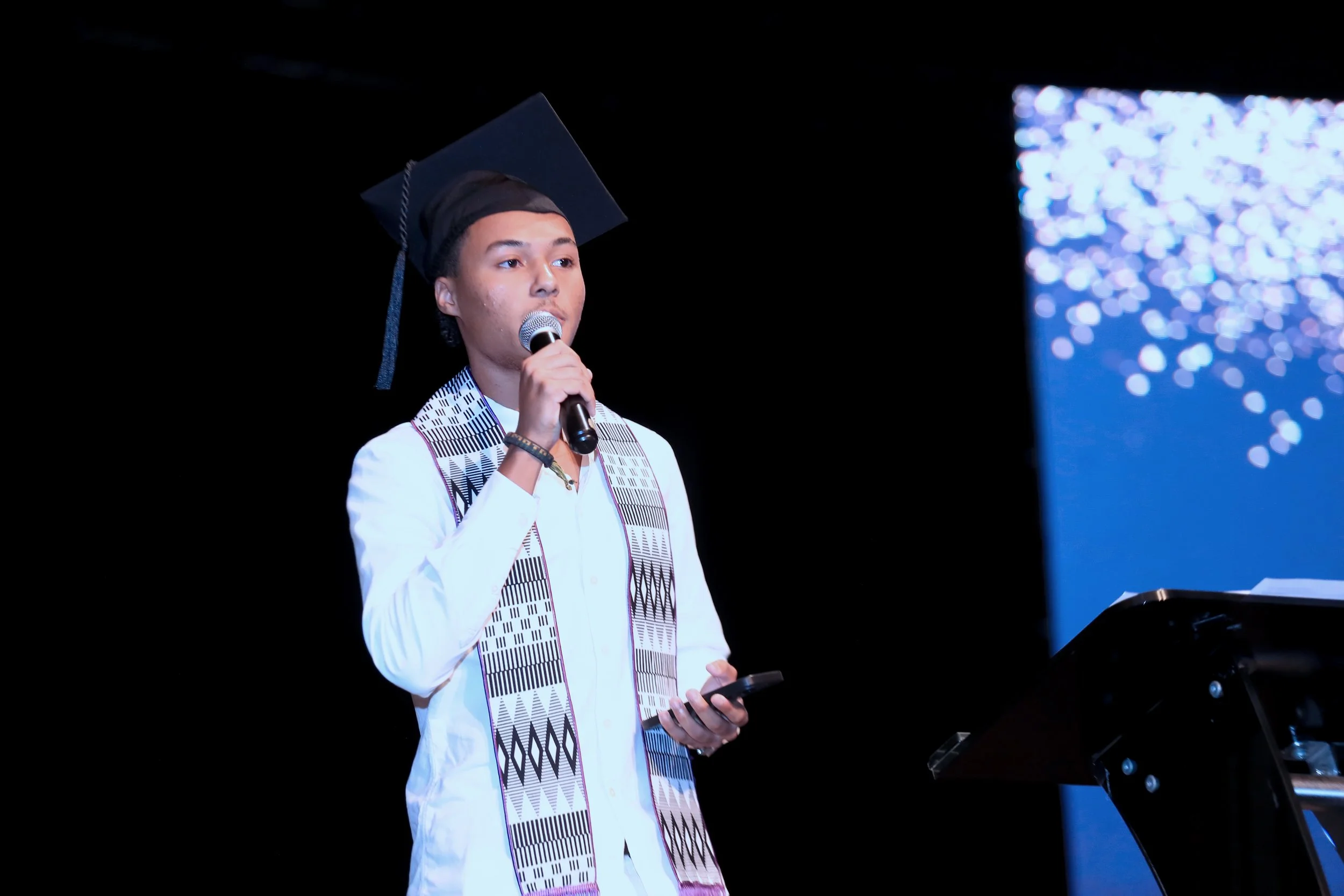 Un jeune homme portant une toge de diplômé, tenant un micro et un téléphone, parlant sur scène lors d'une cérémonie de remise de diplôme, avec un fond sombre et un écran lumineux à droite.