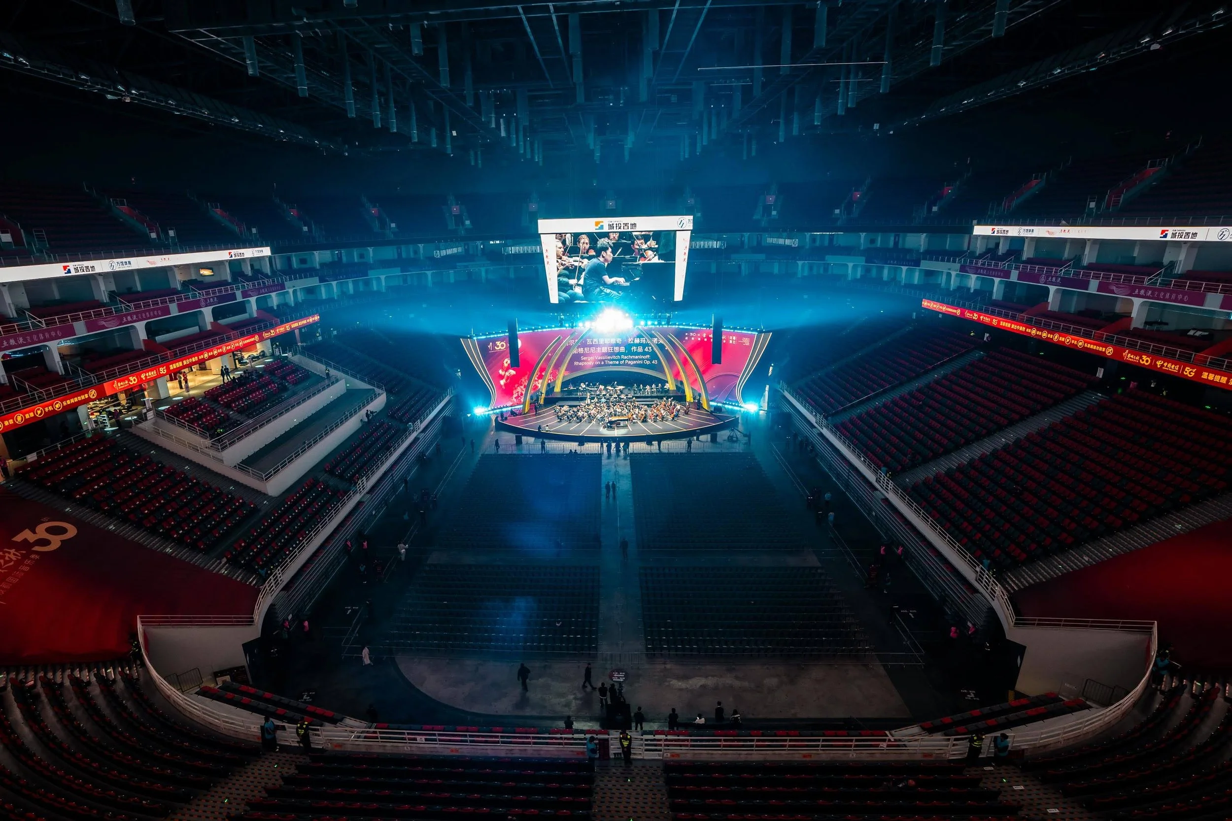 An empty indoor arena with red seats, a stage with an orchestra, and a large screen showing musicians performing, illuminated with bright blue and yellow lights.