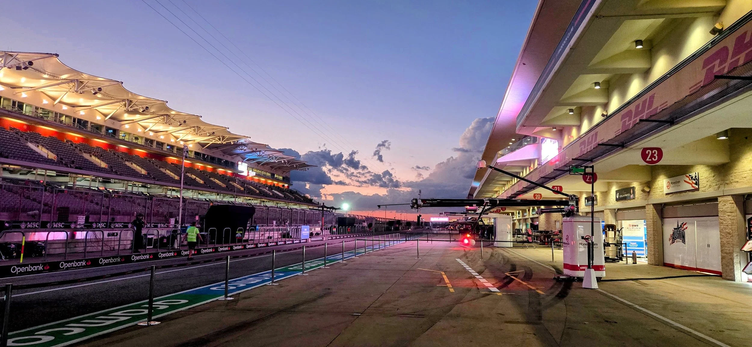 Empty race track paddock with grandstands and team garages at sunset, clouds in the sky, and electronic timing screens
