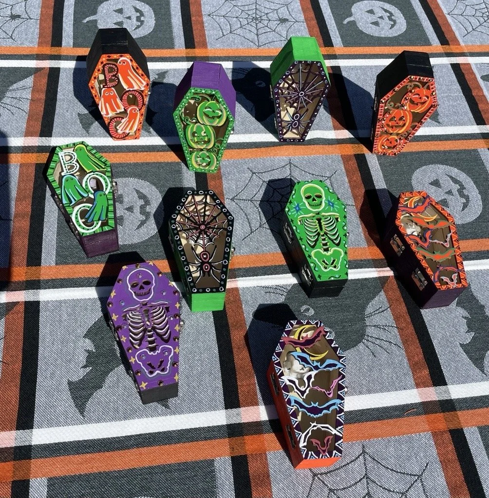 Halloween-themed candy boxes shaped like coffins with colorful spooky and skeleton designs, arranged on a tablecloth with pumpkin and spiderweb patterns.