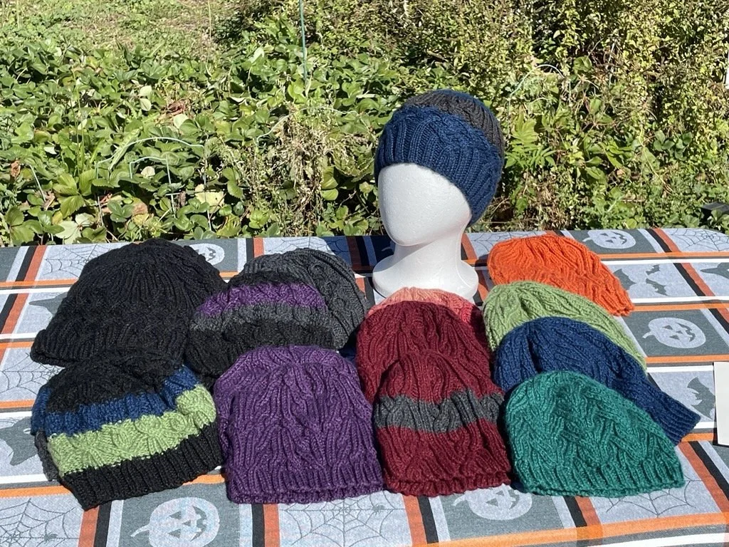 Display of nine knitted beanies on a table outdoors, with one beanie on a mannequin head. The beanies are in various colors including black, purple, green, blue, orange, red, and gray, with different knit patterns.