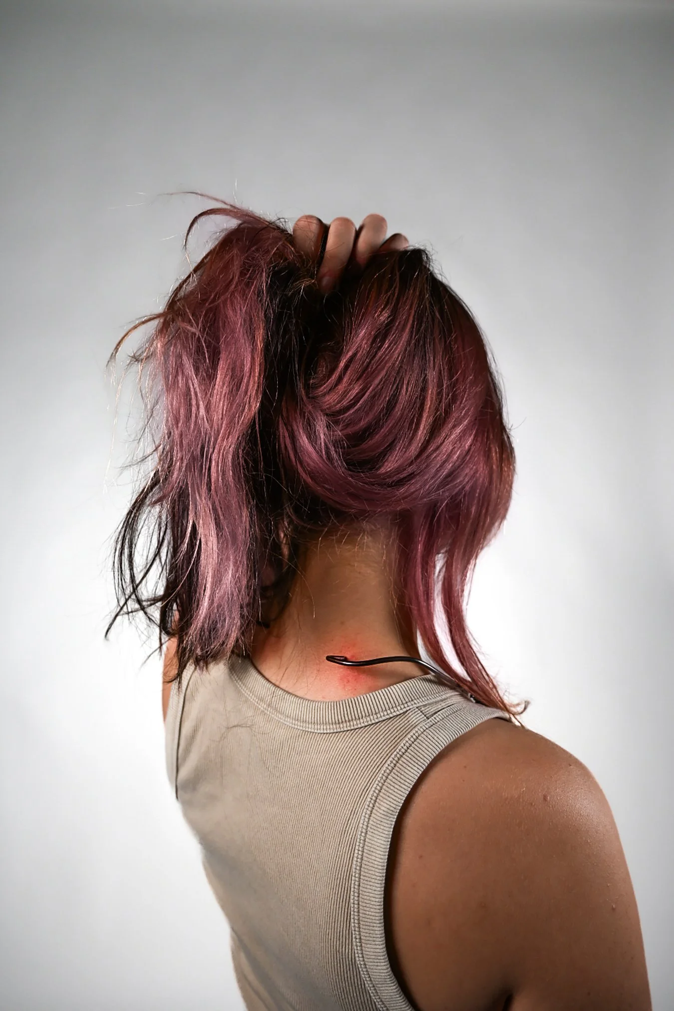A person with short, wavy hair colored with shades of pink and purple, holding their hair up with one hand, wearing a beige sleeveless top, viewed from the back against a plain light gray background.