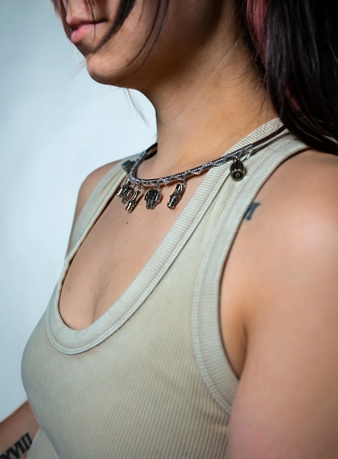 Close-up of a woman wearing a beige tank top and a silver necklace with small charms, showing part of her neck, shoulder, and tattoos.