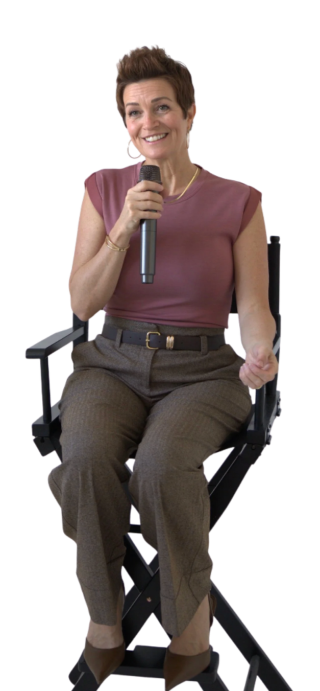 A woman sitting on a director's chair, holding a microphone, smiling, wearing a short-sleeved mauve top, brown trousers, and pointing with her right hand.