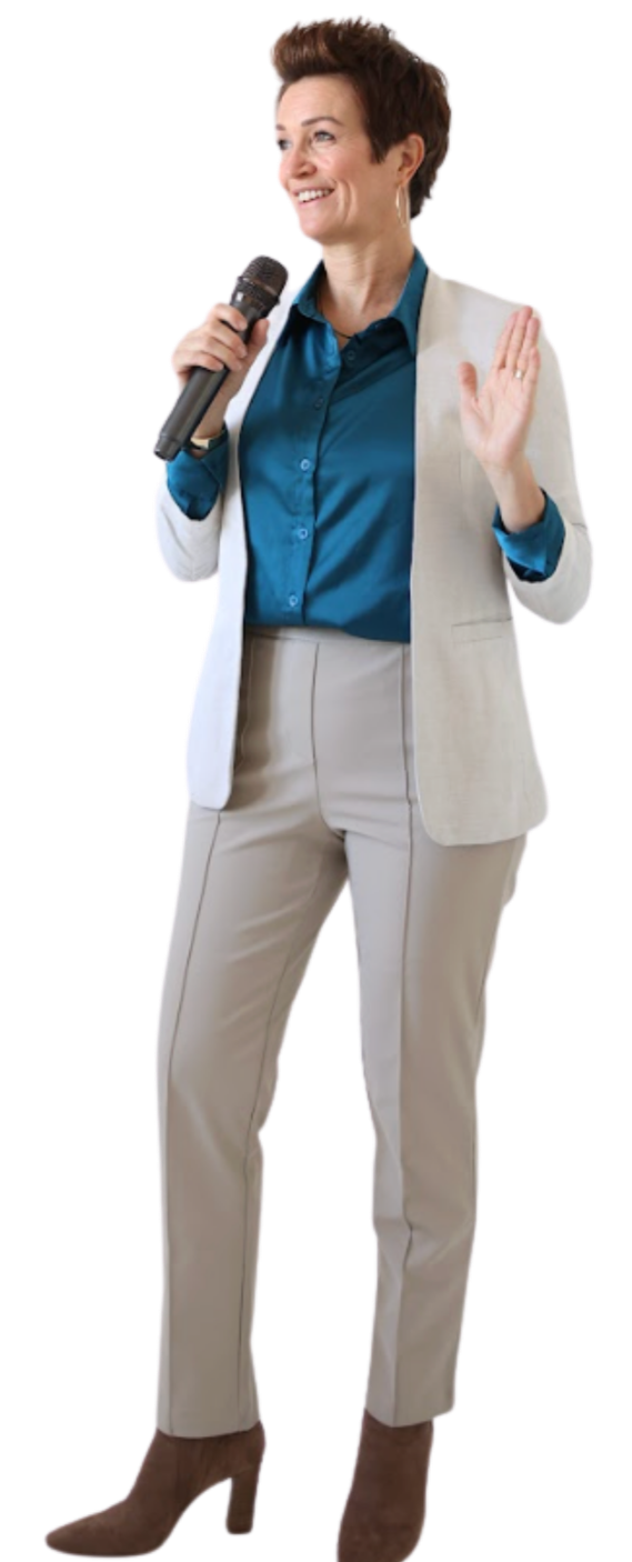 A woman with short brown hair, wearing a blue button-up shirt, a beige blazer, matching beige pants, and brown high-heeled boots, speaking into a microphone and gesturing with her right hand.