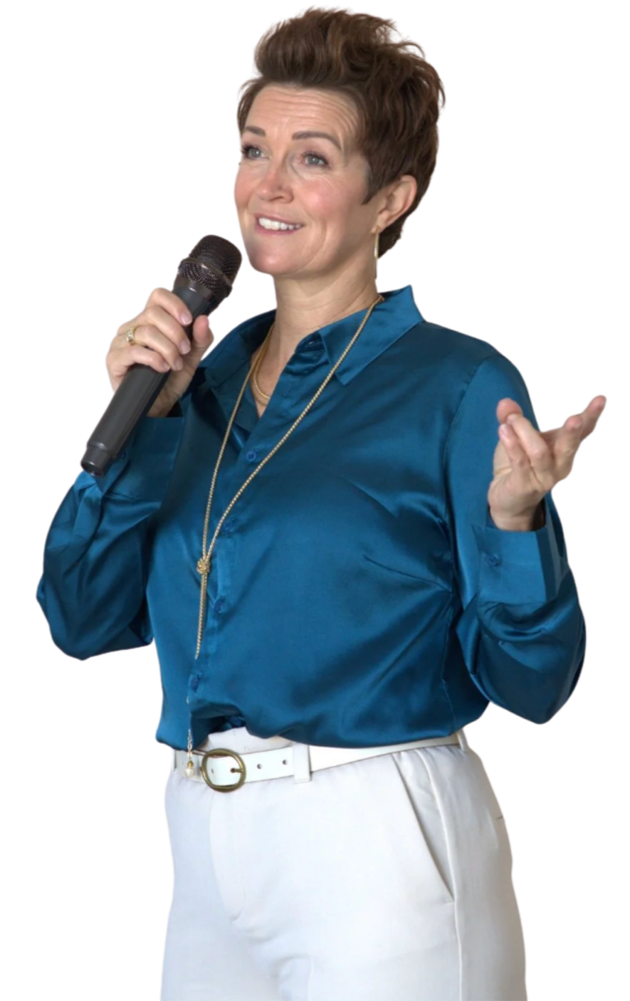A woman with short brown hair, wearing a teal satin blouse and white pants, holding a microphone and gesturing with her left hand.