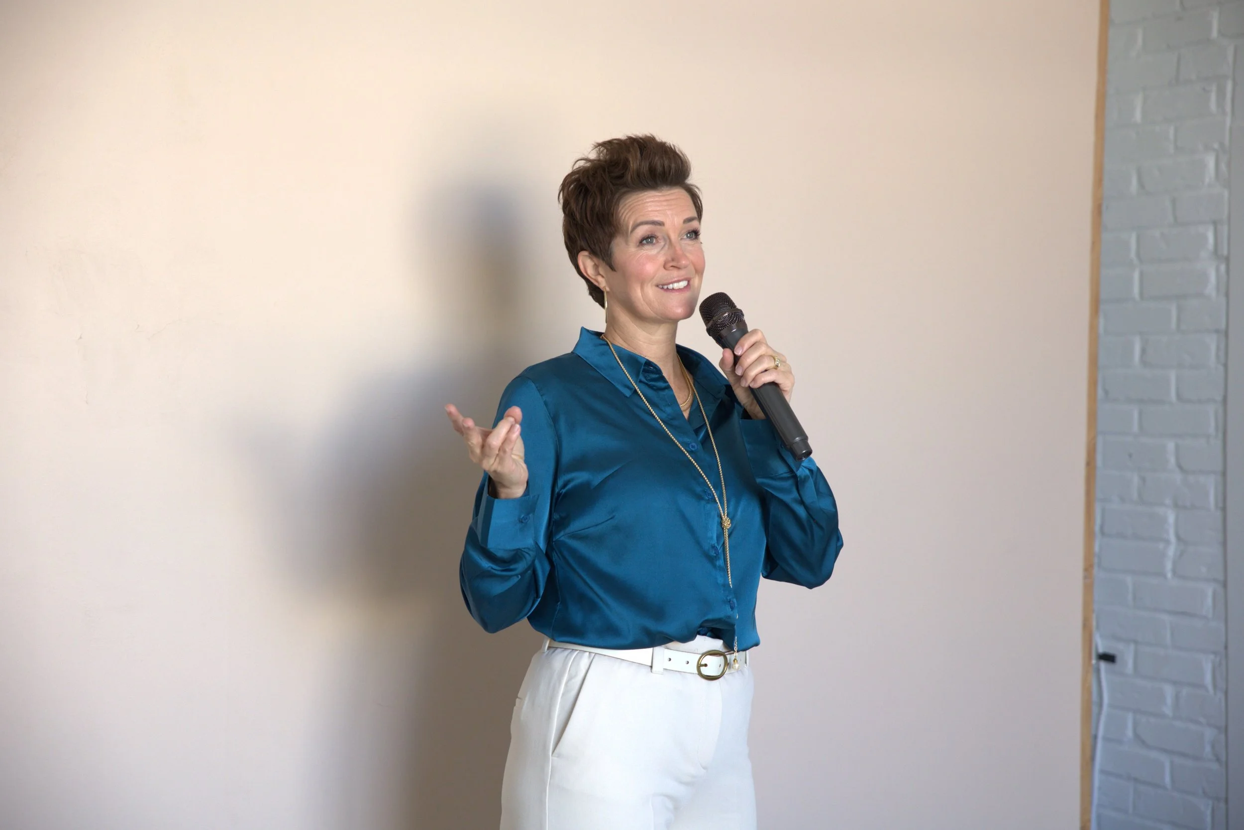 A woman with short brown hair wearing a blue blouse and white pants holding a microphone while speaking or presenting.