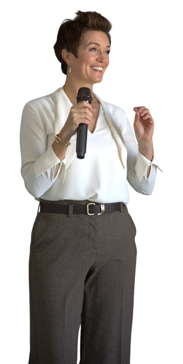 A woman speaking into a microphone, smiling, wearing a white blouse and gray pants.