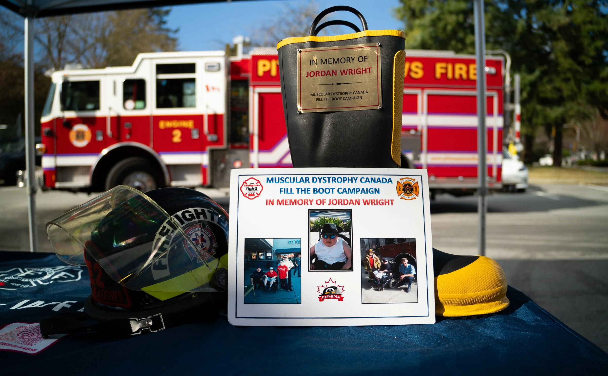 A memorial display featuring a firefighter's helmet, a miniature firefighter's helmet, and a sign dedicated to Jordan Wright, supporting muscular dystrophy awareness and the 'Fill the Boot' campaign, with a fire truck in the background.